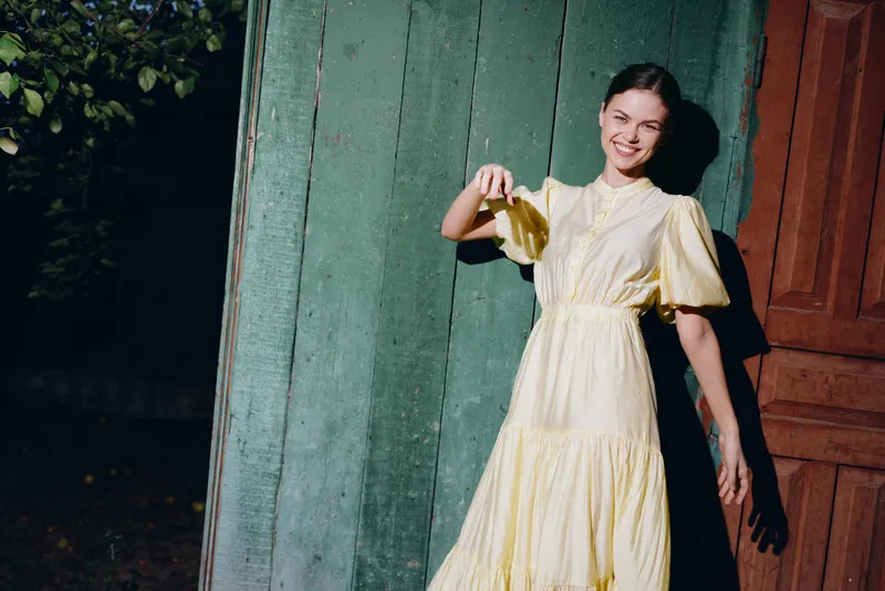 Cheerful woman in a flowing yellow dress smiles as she poses beside colorful doors in an outdoor setting with warm sunlight creating a vibrant artistic mood