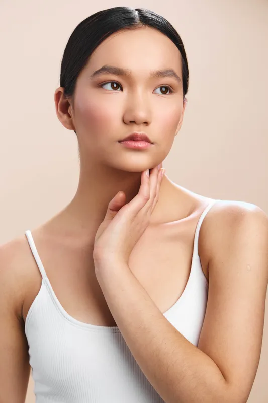 asian woman with calm expression poses in a studio portrait. she wears a white tank top and gentle lighting highlights smooth skin, creating a serene, graceful atmosphere.