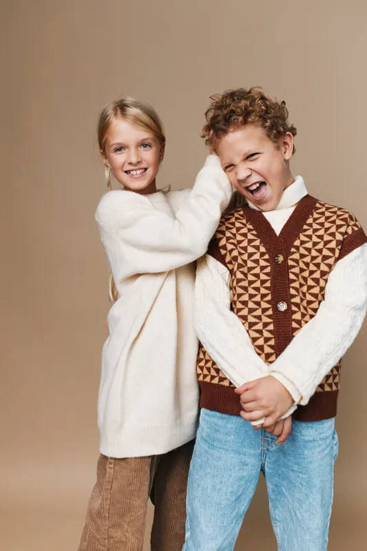 Two cheerful children pose playfully in cozy knitwear, one tugging the other's sleeve. Bright smiles, warm tones, and soft lighting create a friendly, inviting portrait.