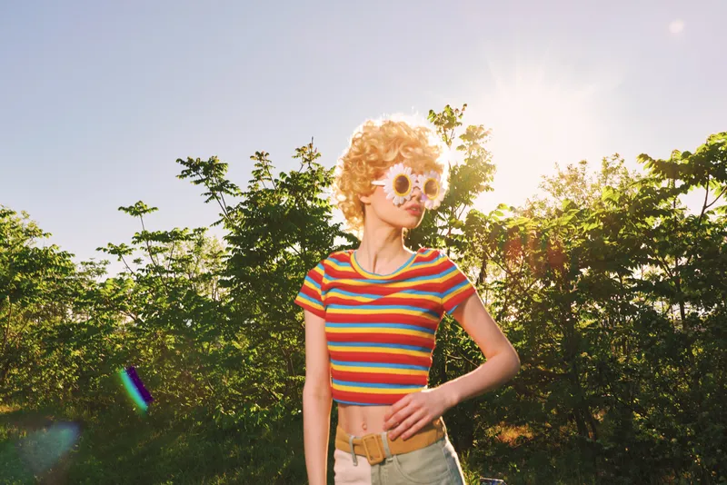 Cheerful woman outdoors wearing a striped crop top and flower sunglasses, sunlit moment with soft breeze, retro film color vibe, natural colors background