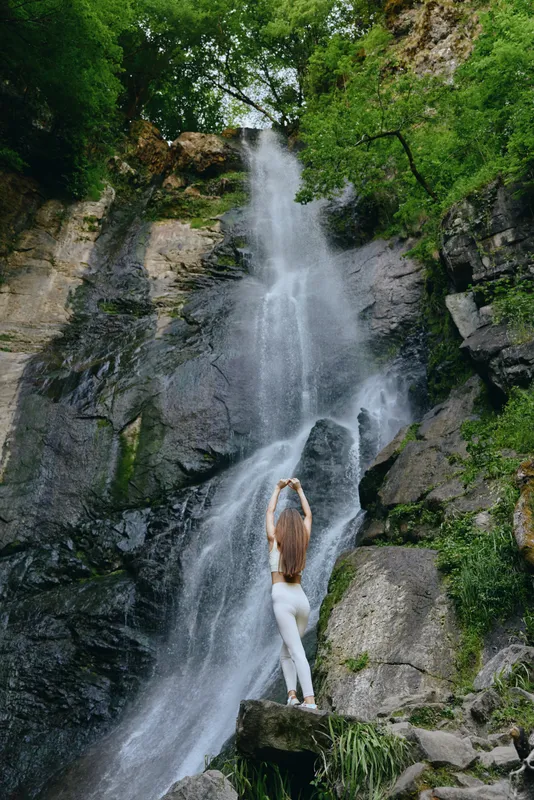 woman stretching near waterfall, natural outdoor scenery, green forest background, rocky cliff, peaceful atmosphere in summer nature setting
