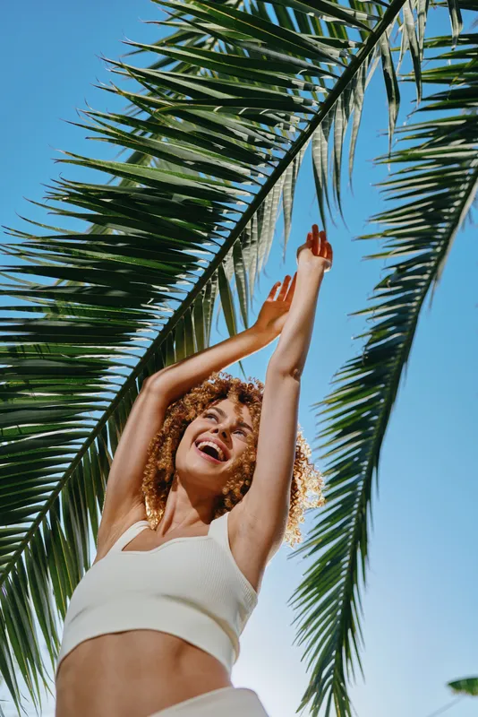 Smiling woman raising hands under palm tree with bright blue sky, curly hair, white crop top, joyful mood in sunny tropical outdoor setting during daytime.