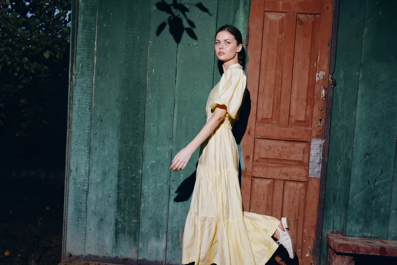 Woman in a flowing yellow dress walks past a green wall and wooden doorway, creating a moody outdoor fashion moment with soft evening light and graceful movement