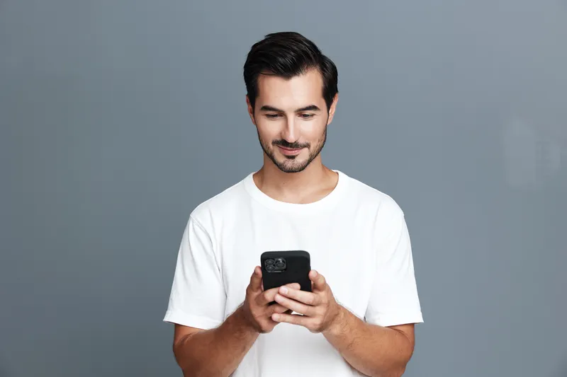 Smiling young man wearing a casual white t-shirt, looking at a smartphone while standing against a plain grey background. Modern technology and lifestyle concept.