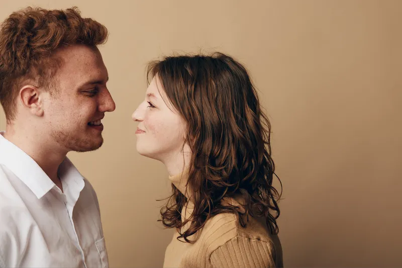 Young couple with red hair and brown hair face each other closely, smiling gently. Intimate moment captured on beige background. Concept of love, connection, and affection.