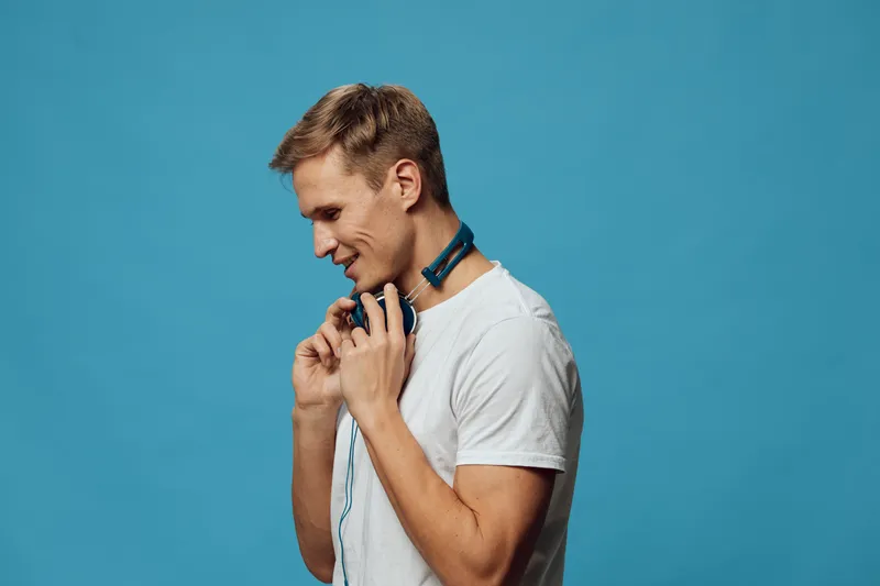 Smiling young man wearing casual white t-shirt listening to headphones against blue background, enjoying music, leisure, relaxation concept, studio portrait, lifestyle.