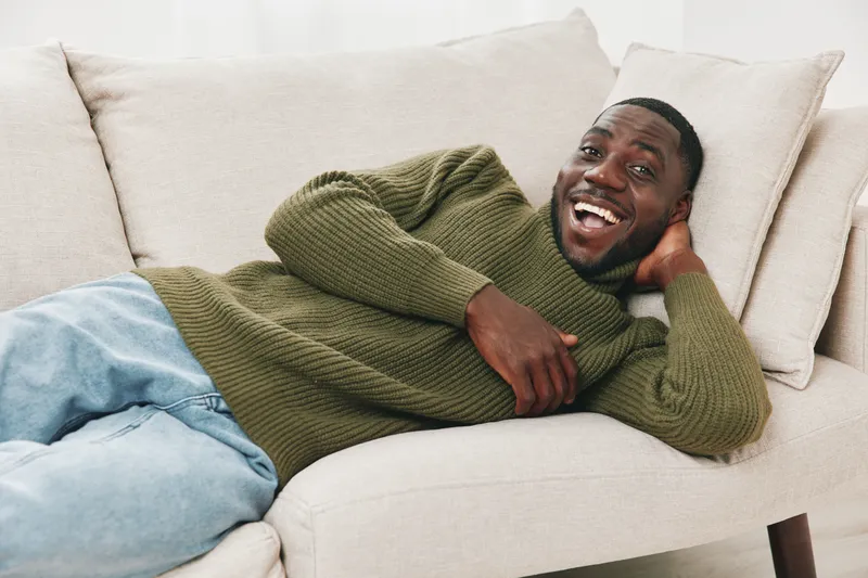 A joyful young African man lounging on a cozy couch, wearing a green sweater and blue jeans, radiating happiness and comfort in a bright, neutral toned living room