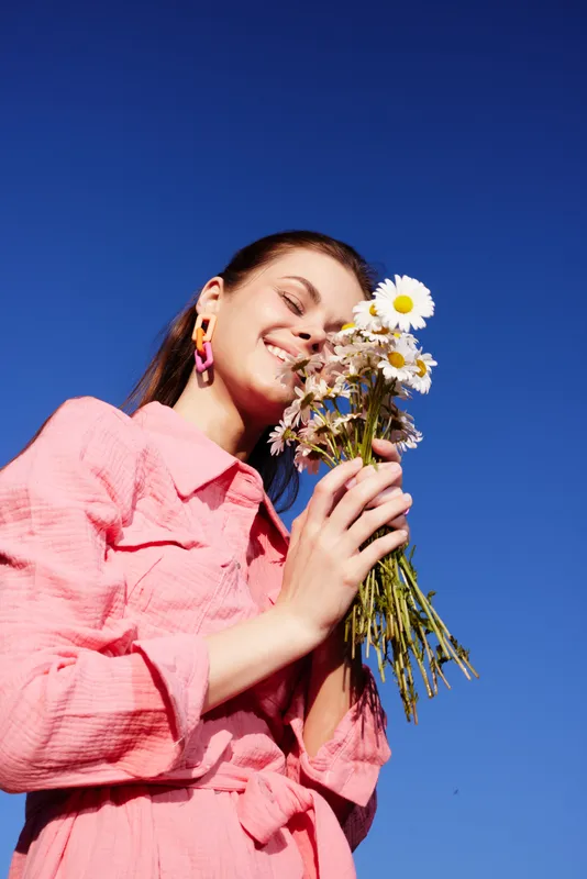 Joyful young woman in a pink dress holding daisies against a bright blue sky, embodying a cheerful and vibrant spring moment