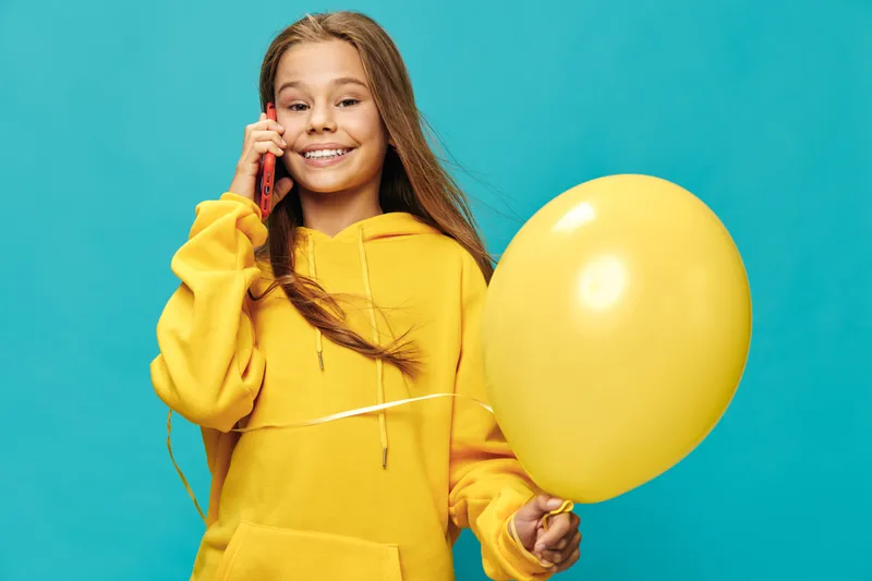 Girl in yellow hoodie holding yellow balloon talking on smartphone smiling on blue background. Teenager, happy child and communication in casual clothing with long hair smiling indoors.