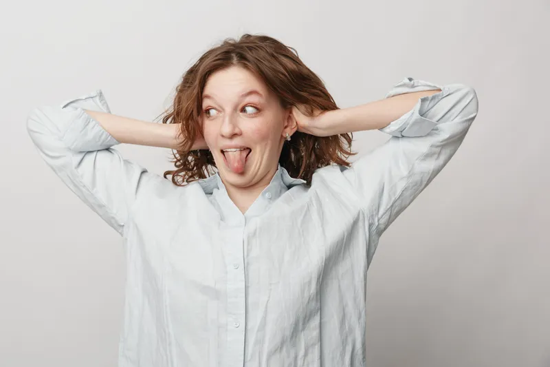 Playful young woman with curly hair in white shirt makes funny face sticking out tongue and holding hands behind head. Studio portrait on light background with casual mood and expression.
