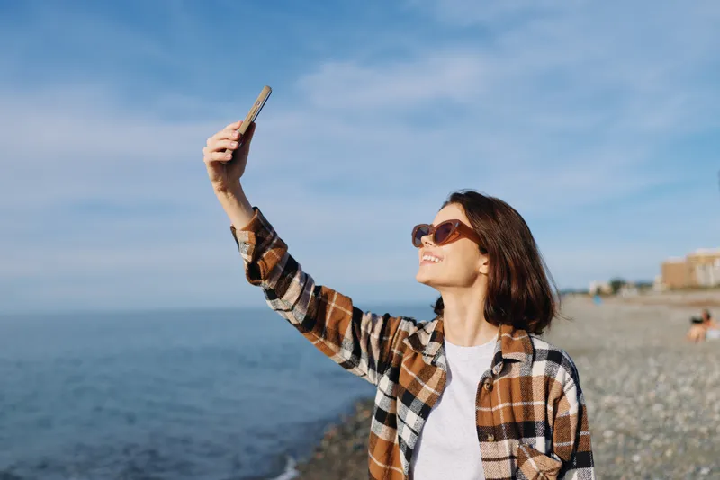 Selfie of a woman by the ocean wearing a plaid shirt and sunglasses, capturing a breezy coastline moment with phone held aloft against a clear sky, casual style and sunny atmosphere.
