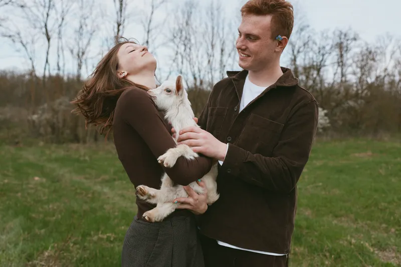 Joyful couple outdoors holding a white goat kid, laughter and affection in a park setting, casual autumn attire, soft natural light, candid lifestyle moment