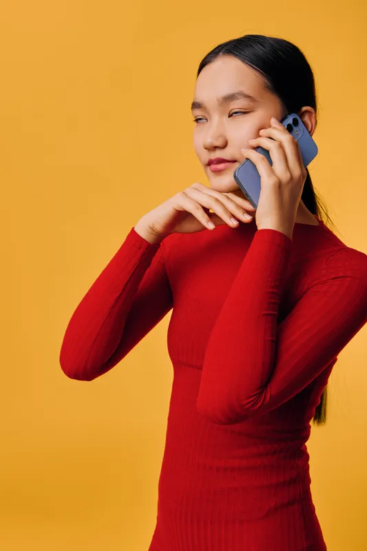 Young woman in red dress talking on smartphone with thoughtful expression posing on yellow background. Portrait of female holding mobile phone close to face with minimalistic studio color.