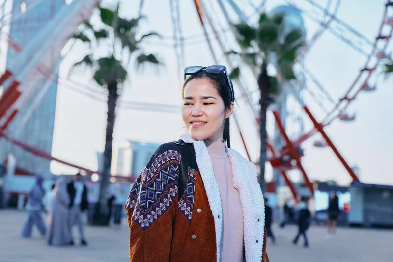 Woman in a knitted cardigan and scarf strolls through an outdoor amusement park with a ferris wheel in the background, sunny day, candid fashion pose, casual travel vibe.