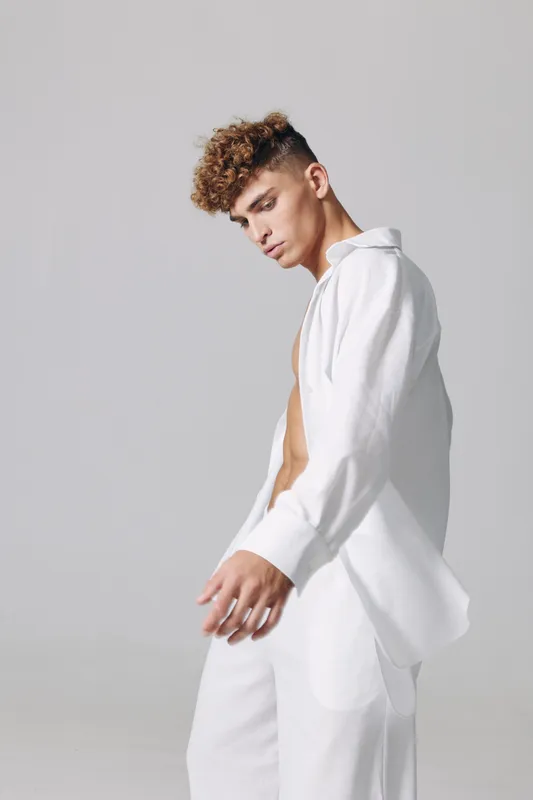 Young man with curly hair wearing white shirt and pants posing in studio with neutral background. Fashion model with casual style and thoughtful expression in minimalistic setting.