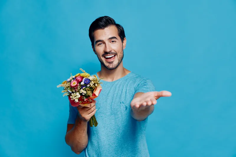 Happy smiling man in casual blue t-shirt holding a bouquet of colorful flowers with a joyful expression, looking directly at the camera against a bright blue background.