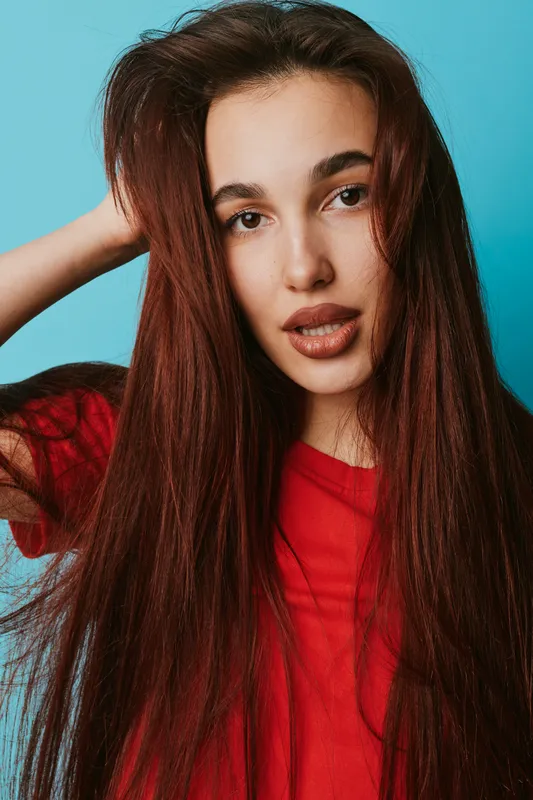 Young woman with long straight hair wearing red shirt poses with hand in hair against blue background. Portrait of female model with natural makeup and confident expression.