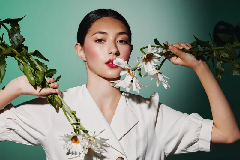 Young creative woman in white blouse posing with large white flowers against a soft green background. Bold makeup enhances her confident and stylish beauty concept.