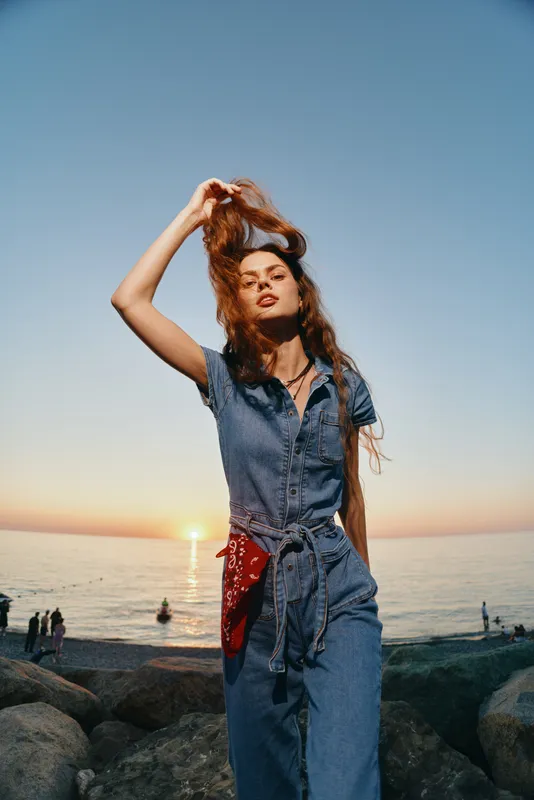 young woman in denim jumpsuit standing on rocky beach during sunset with relaxed, confident pose, natural light, and serene ocean background