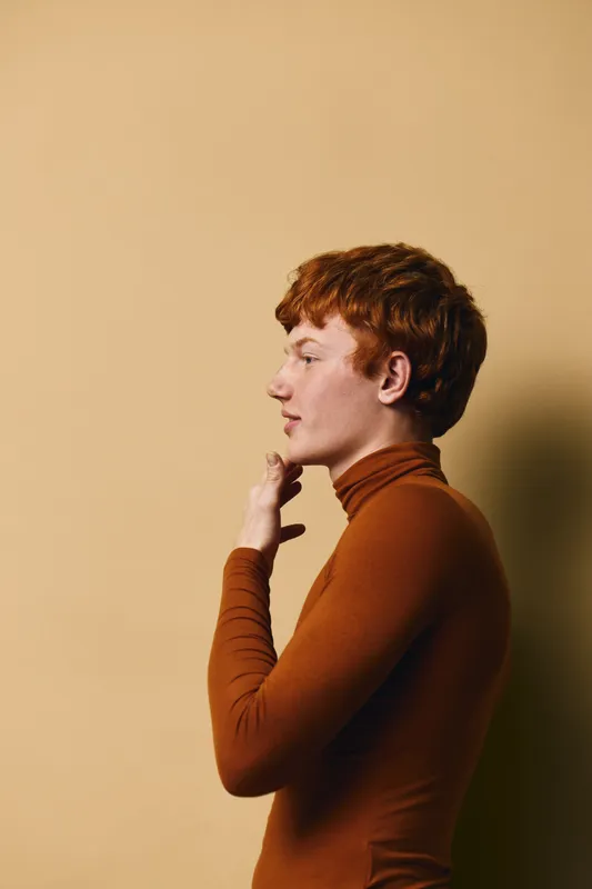 Profile portrait of redhead young man wearing brown turtleneck sweater standing against beige background with thoughtful expression and hand near chin.