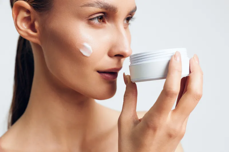 Young woman with long brunette hair applies white face cream on her cheek while holding an open jar in a bright studio setting focused on skincare and self-care routines.