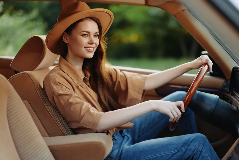 A cheerful young woman with long hair wears a stylish hat and a brown shirt while driving a car, surrounded by a vibrant green background.
