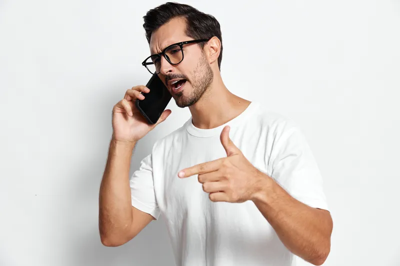 Energetic man talking on mobile phone, wearing glasses and white t-shirt, gesturing dramatically while engaging in conversation, isolated on plain white background.