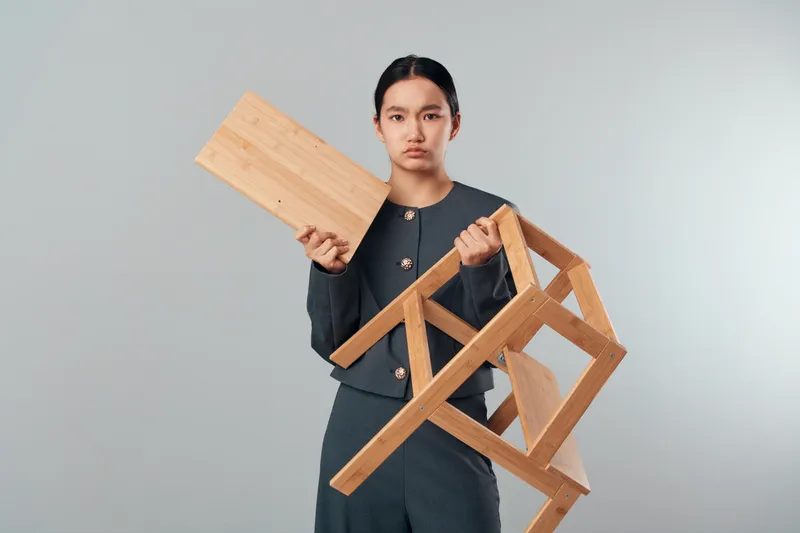 Young woman holding a broken wooden chair and a detached chair seat, serious expression and plain background for furniture repair or woodworking concept.