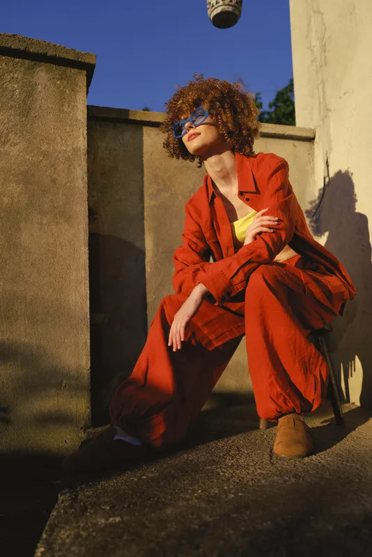 A woman in a bold red suit crouches on a sunlit rooftop, wearing sunglasses and posing with confidence. An urban outdoor scene highlighting fashion, lifestyle, and creative street style