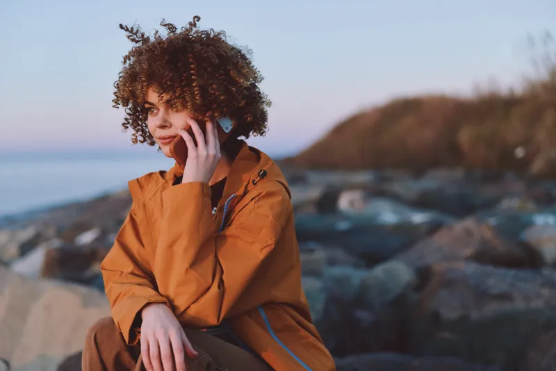 Woman lifestyle outdoors by sea and ocean at dusk, curly hair, orange jacket, sitting on rocks holding a phone to her ear, calm expression, seaside