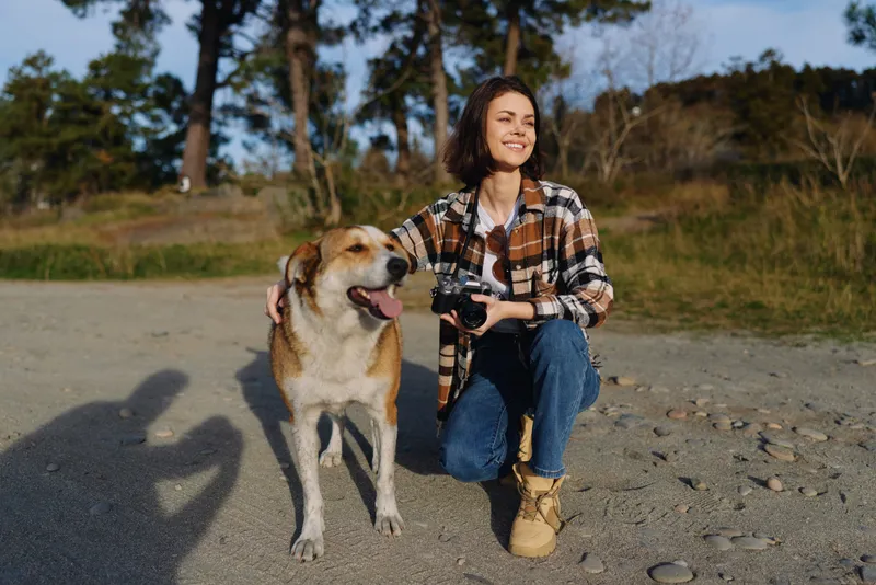 Casual outdoor portrait of a smiling woman kneeling beside a friendly dog while holding a camera, in a natural park setting with trees and sunlight, capturing a moment of companionship