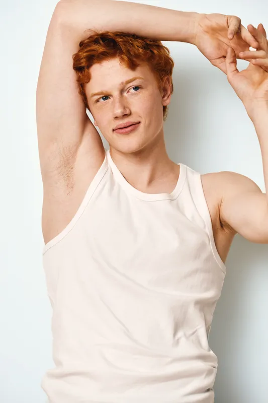 Young man with red hair in white tank top stretching arms above head with relaxed face against plain background. Casual style, natural pose and confident expression.