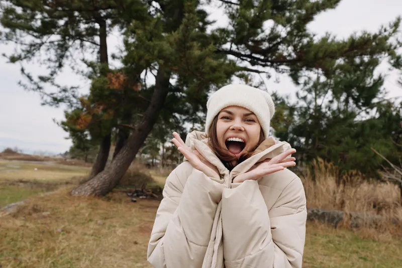 Woman outdoor winter coat joyful expression, wearing beanie and scarf, standing near trees in a windy field, candid lifestyle shot in natural light