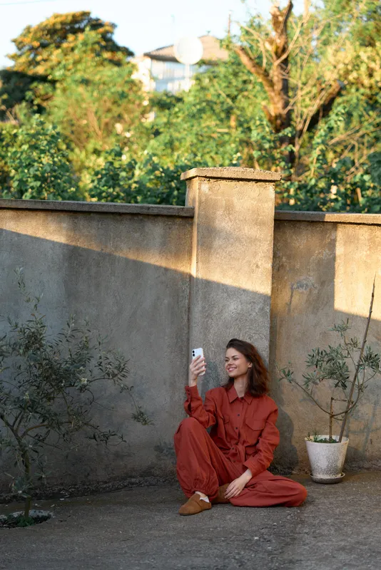 A woman in a rust red jumpsuit sits on a concrete courtyard taking a selfie with her phone, sunlight over a weathered wall and potted plants creating a casual lifestyle moment