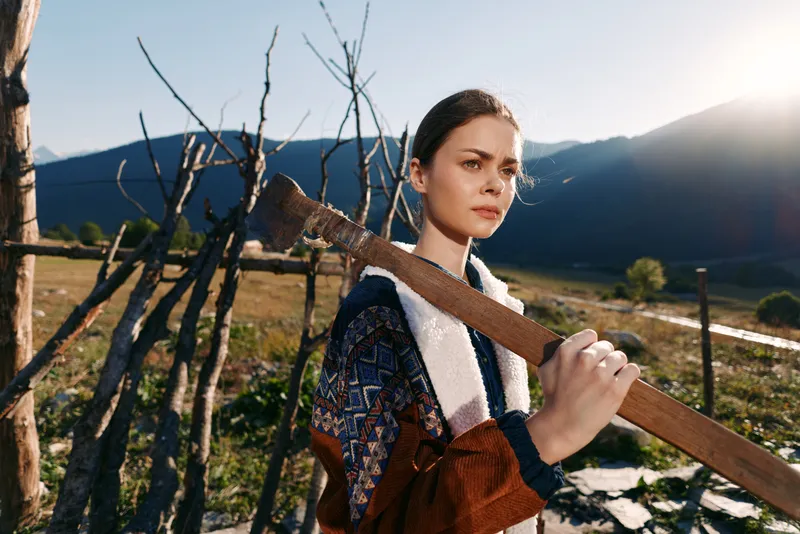 Woman carrying wooden plank near rustic fence with mountains in background, rural outdoors portrait at sunrise, young female in patterned jacket holding timber in scenic landscape.