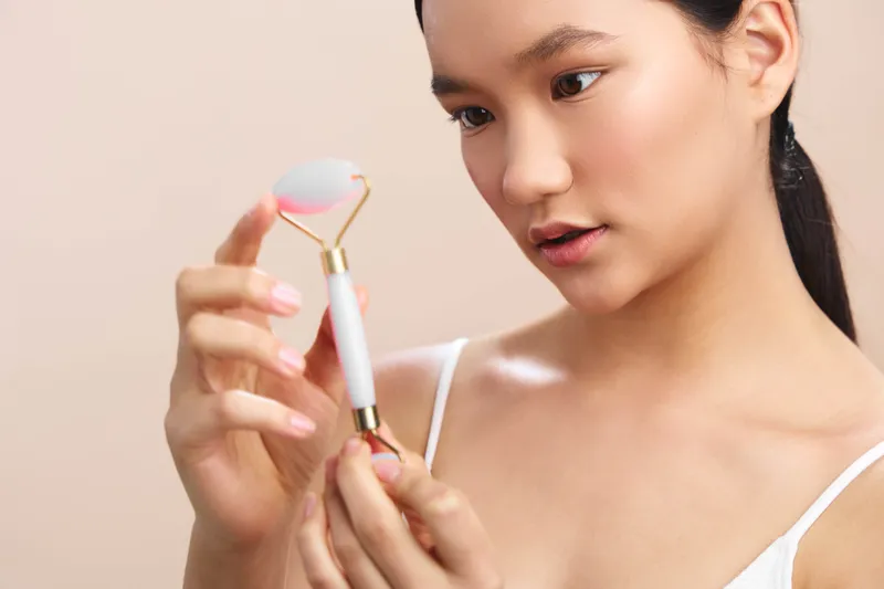 asian woman is seen using a facial roller in a soft neutral setting, focused and calm, illustrating a skincare routine that promotes healthy glow and mindful selfcare.