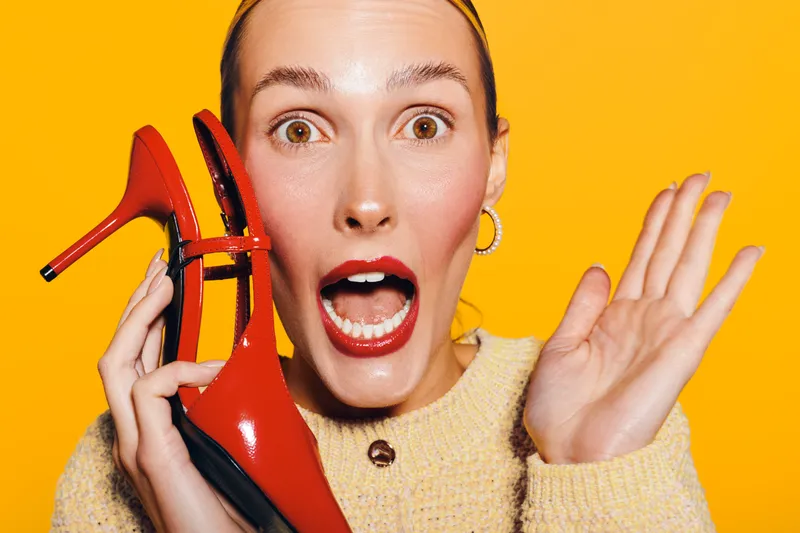 Surprised woman with red lipstick holds red high heel shoe near face on bright yellow background. Concept for fashion, beauty, and emotional expression in studio portrait.