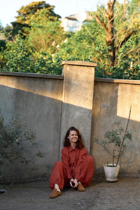 Casual outdoor portrait of a woman in orange attire, relaxed posture, sunlit moment among greenery, candid lifestyle scene highlighting everyday confidence and natural colors