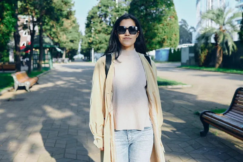 Woman wearing a beige coat and knit sweater, sunglasses, strolling along a sunny park street with trees and benches, capturing casual fashion and outdoor lifestyle, bright atmosphere, relaxed