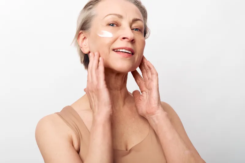 Mature woman with short hair applies moisturizing cream on her face in a bright studio. Concept of skincare, aging beauty, self-care, and healthy skin routine for older adults.