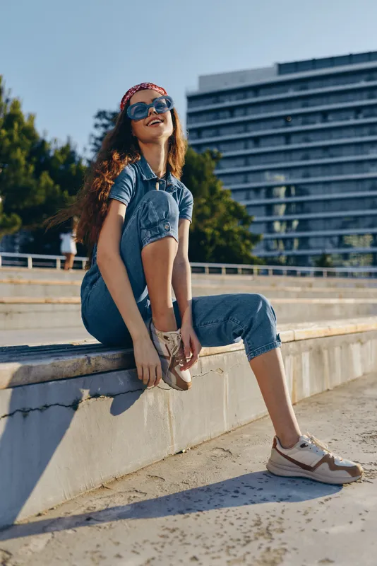 smiling woman sitting outdoors in casual denim overalls and sunglasses, enjoying sunny day with relaxed and happy mood in urban environment