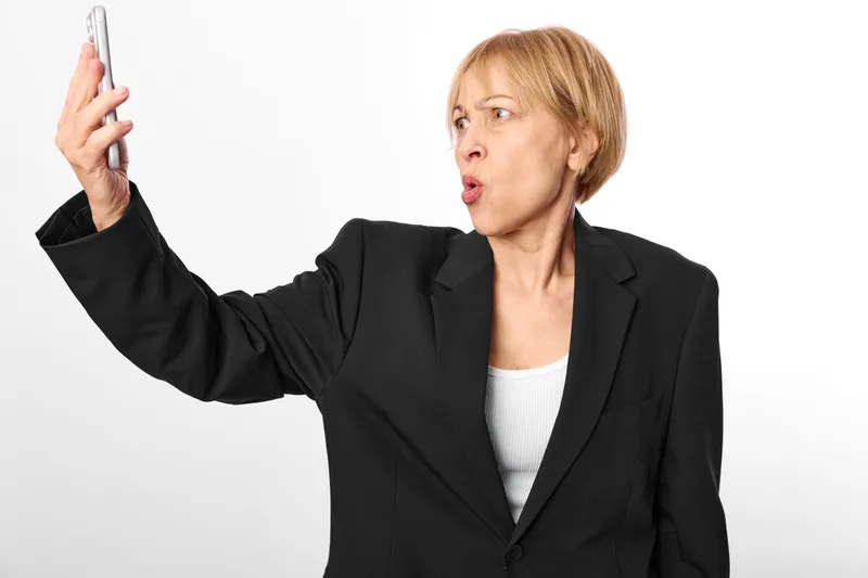 Shocked woman holding smartphone with raised gesture in black blazer on bright white background, surprised business communication, studio portrait, copy space