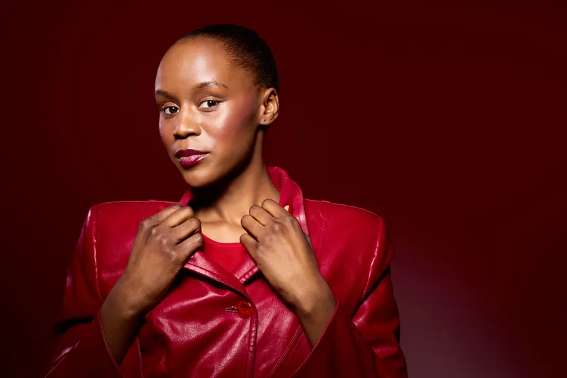 Confident dark skinned African woman wearing a red leather jacket poses against a red background. Studio portrait with strong lighting highlights facial features and stylish clothing.