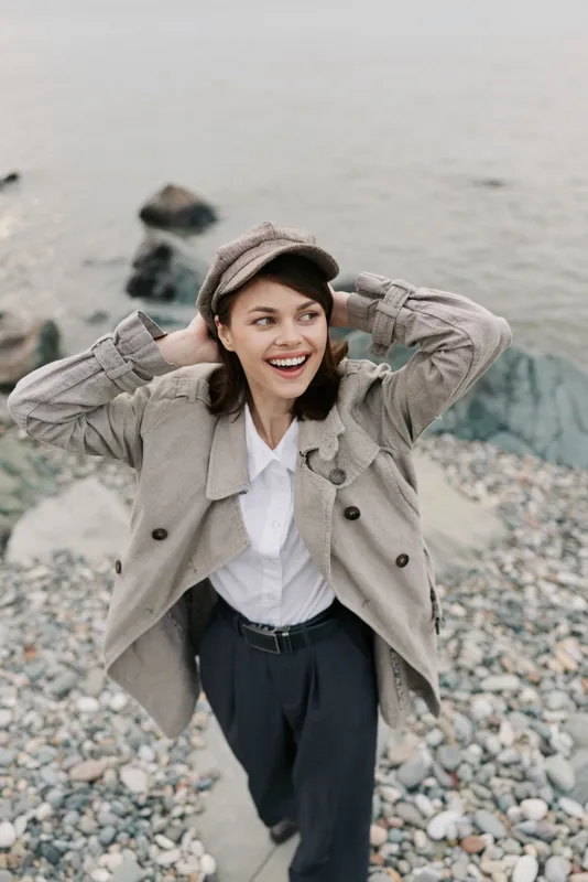 Happy young woman in casual autumn outfit stands on rocky beach near water smiling joyfully. She wears a textured coat and a hat, enjoying fresh air and nature outdoors in cool weather.