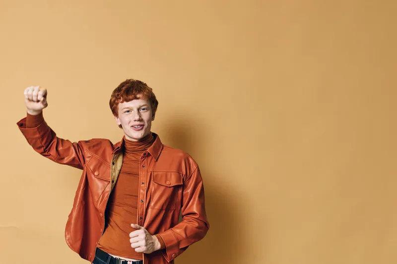 Young man with red hair wearing orange jacket and brown turtleneck celebrating success with raised fist and smiling on plain beige background.