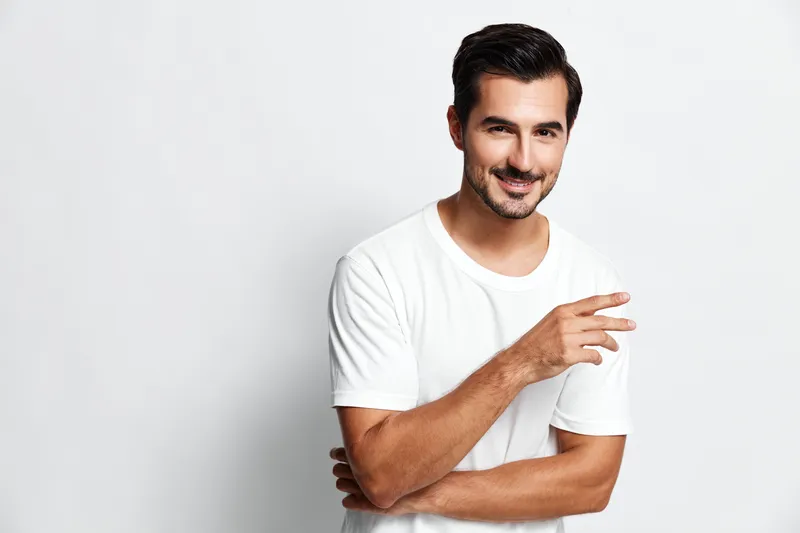 Smiling man wearing a white t-shirt poses confidently with a friendly gesture against a simple light background, conveying warmth and approachability.