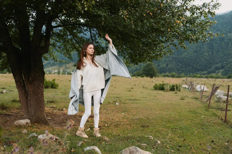 A young woman dances in a breezy meadow, wearing white pants and a beige knit sweater with a gray shawl flowing, mountains in the background
