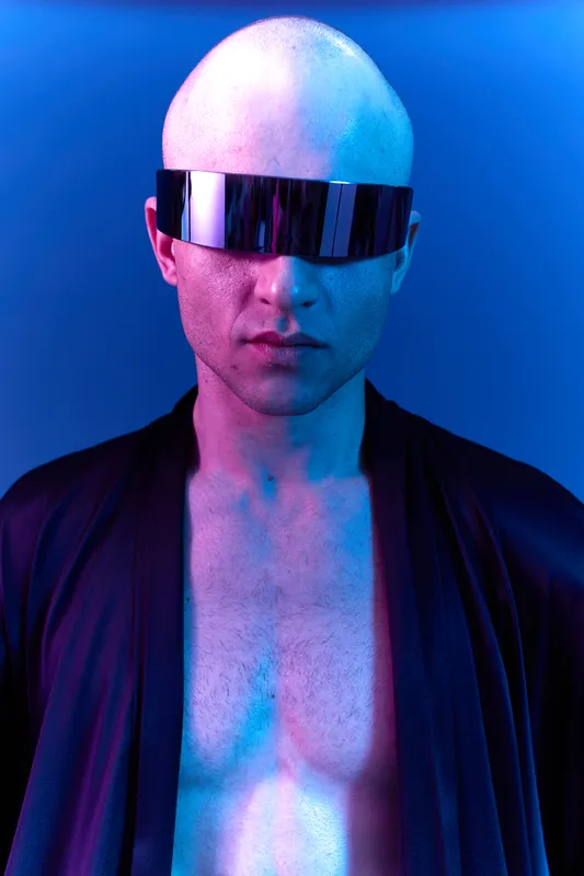 A striking man with a bald head and futuristic sunglasses displays confidence against a vivid blue background. The image radiates mystery and modern style.