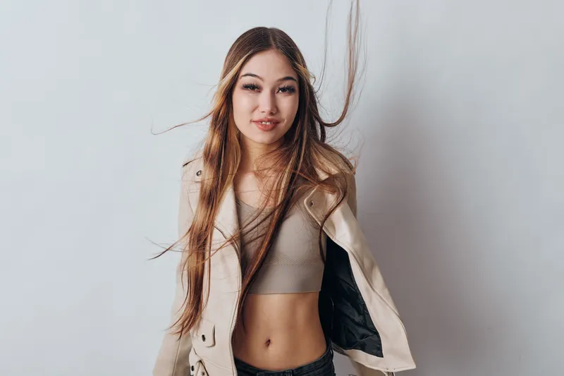 Young woman with long hair wearing casual top and open jacket posing confidently against plain light background. Beautiful female model with natural makeup and relaxed style in studio portrait.