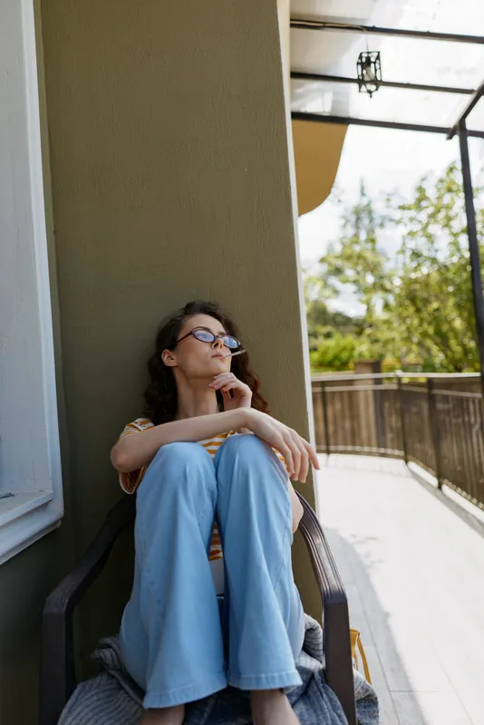 Woman relaxing outdoors at home on a balcony, seated in a chair, wearing light blue jeans and sunglasses, thoughtful pose, sunlit scene, soft breeze, casual modern mood and relaxed posture